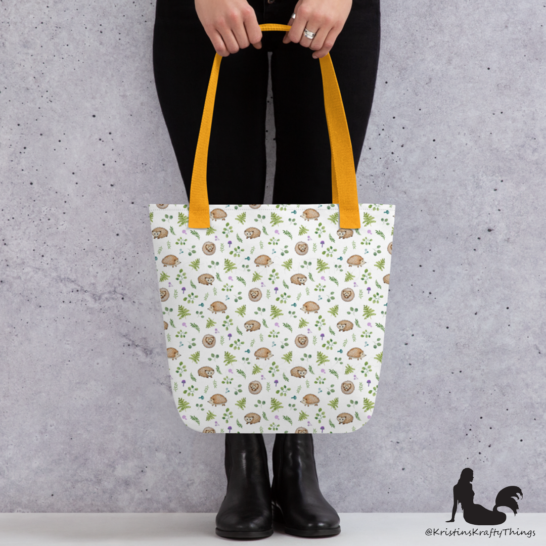 All-over full color print from original watercolor painting. These adorable woodland hedgehogs dance about with green ferns and purple & teal mushrooms on this spacious tote bag to help you carry around everything that matters. Photo of tote bag with yellow handle being held by a person to show size reference.