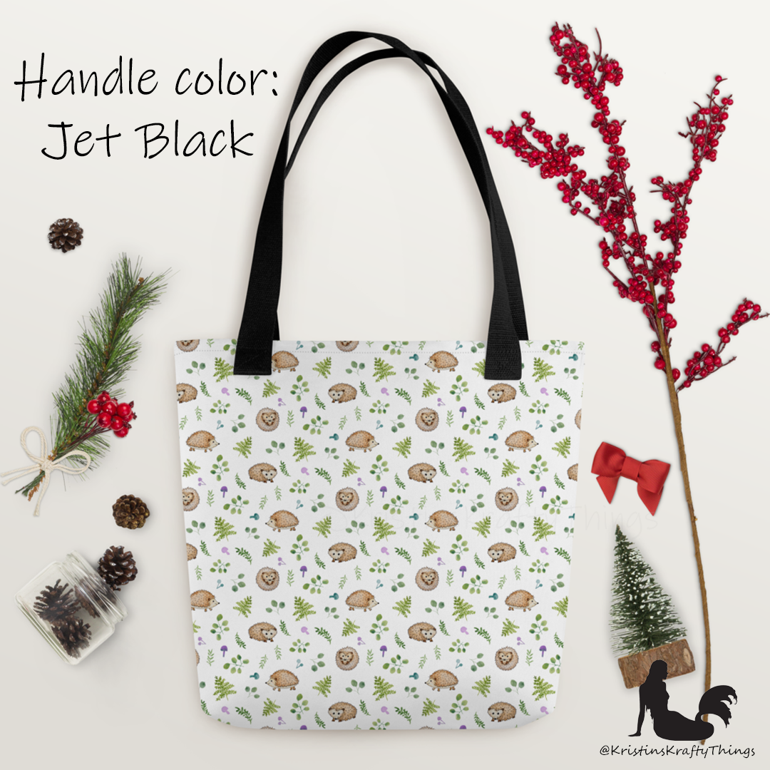 All-over full color print from original watercolor painting. These adorable woodland hedgehogs dance about with green ferns and purple &amp; teal mushrooms on this spacious tote bag to help you carry around everything that matters. Photo of tote bag with black handle on holiday layout.