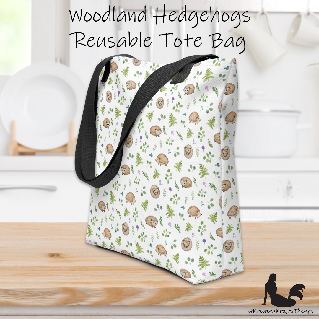 All-over full color print from original watercolor painting. These adorable woodland hedgehogs dance about with green ferns and purple &amp; teal mushrooms on this spacious tote bag to help you carry around everything that matters. Photo of tote bag on counter top.