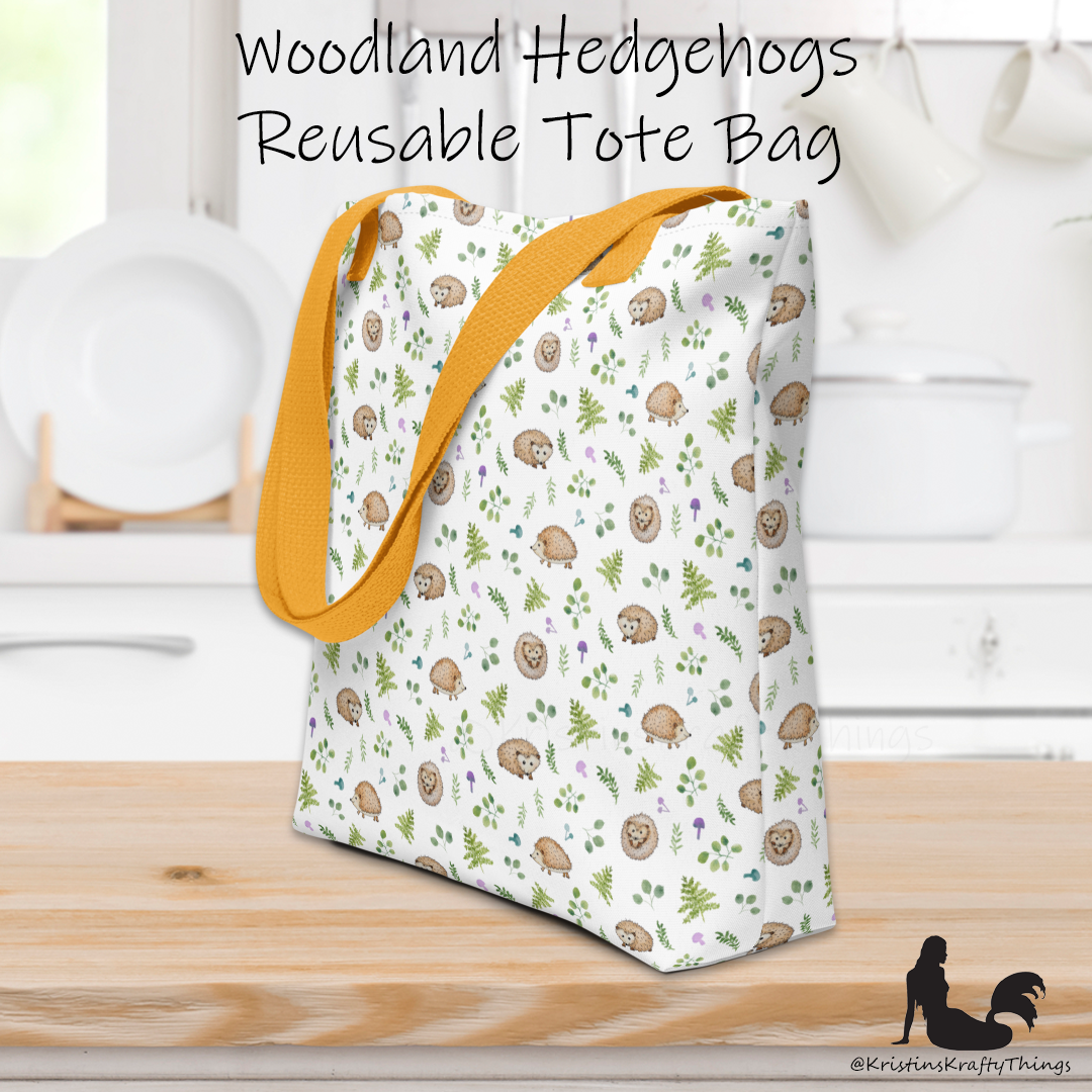 All-over full color print from original watercolor painting. These adorable woodland hedgehogs dance about with green ferns and purple &amp; teal mushrooms on this spacious tote bag to help you carry around everything that matters. Photo of tote bag on counter top.