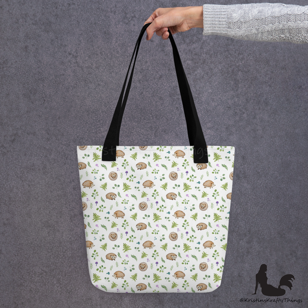 All-over full color print from original watercolor painting. These adorable woodland hedgehogs dance about with green ferns and purple &amp; teal mushrooms on this spacious tote bag to help you carry around everything that matters. Photo of tote bag with black handle being held by an outstretched arm.